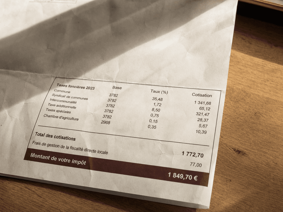 Une feuille d'avis de taxe foncière posée à plat sur une table en bois clair, froissée aux coins comme relue plusieurs fois, éclairée par la lumière rasante d'une fenêtre qui fait ressortir les chiffres imprimés en noir.