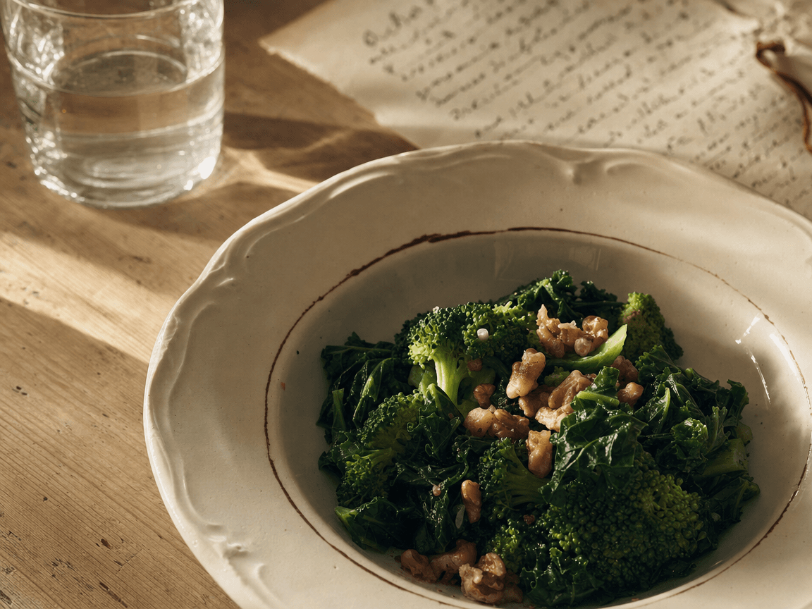 Une assiette creuse en faïence blanche posée sur une table en bois clair, à moitié remplie de légumes verts et de noix, photographiée en légère plongée dans la lumière rasante d'une fin d'après-midi, un verre d'eau et une feuille manuscrite illisible en arrière-plan flou.