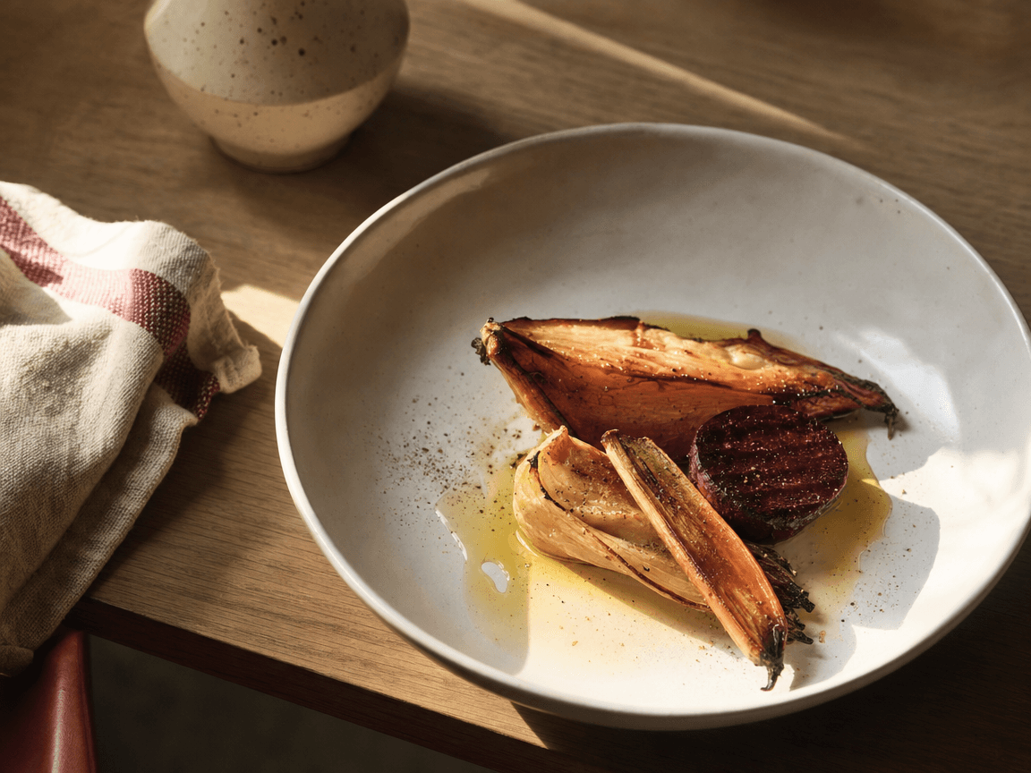 Une assiette creuse en céramique blanche posée sur une table en bois clair, photographiée en plongée légère, où quelques légumes rôtis aux teintes ocre et bordeaux côtoient un filet d'huile d'olive qui capte la lumière de fin d'après-midi.